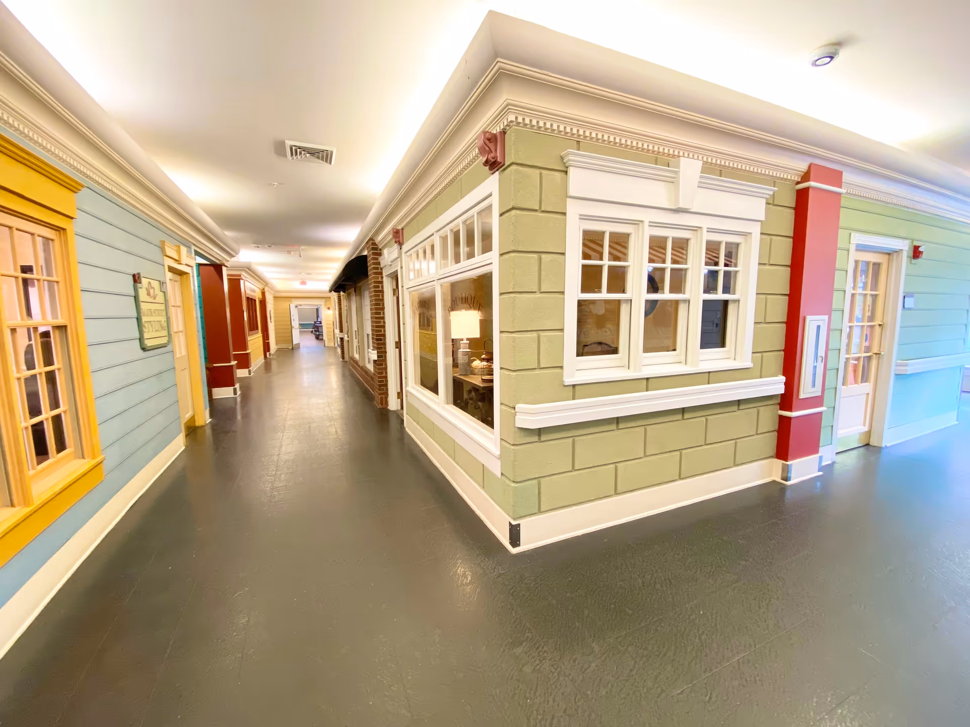 A brightly lit indoor hallway with walls designed to look like the exterior of houses, featuring windows and doors in various colors including yellow, green, and blue. The floor is dark and smooth, and the ceiling has recessed lighting and crown molding.