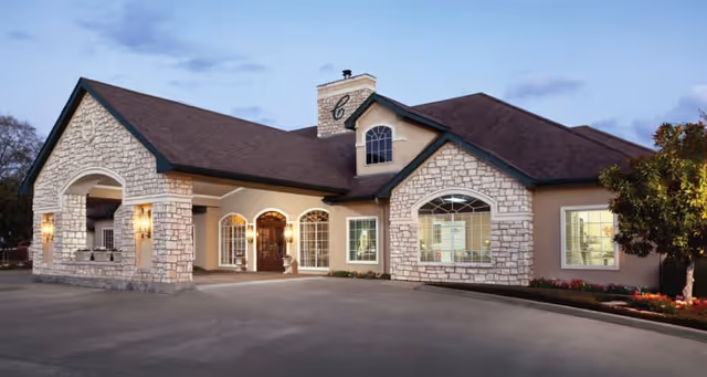 Exterior view of a single-story building with stone and beige stucco walls, a dark brown roof, large windows, and a covered entrance with lights on either side of the door, set against a partly cloudy sky at dusk.