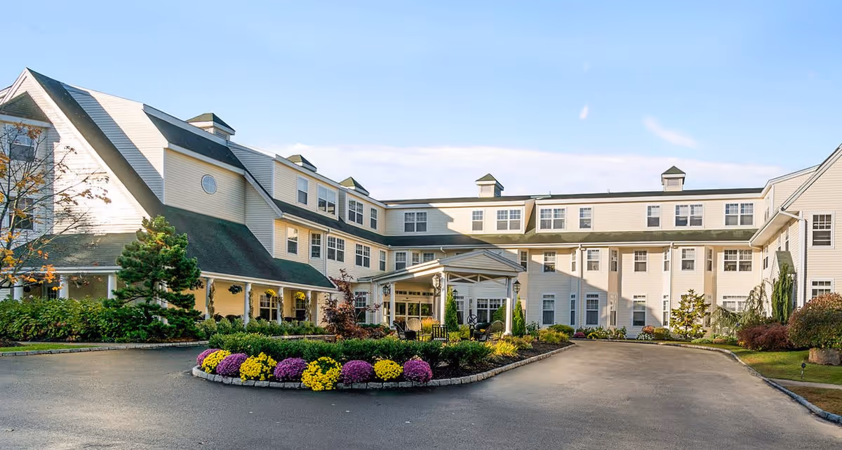 Exterior view of a large, multi-story senior living facility with beige siding and green roofing. The building surrounds a circular driveway with a landscaped island featuring colorful flowers and shrubs. The sky is clear with a few clouds.