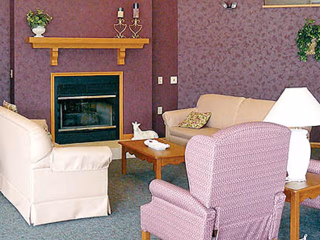 A cozy living room with a beige sofa, a cream armchair, and a purple patterned armchair arranged around a wooden coffee table. The room features a fireplace with a wooden mantel decorated with candles and a small plant. There is a table lamp on a side table next to the purple armchair, and the walls are covered with purple floral wallpaper.