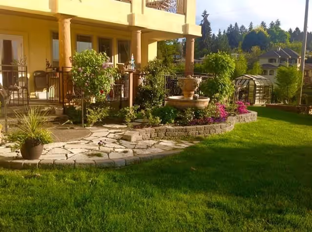 A well-maintained outdoor garden area with a stone pathway, green grass, potted plants, a raised flower bed with colorful flowers, and a water fountain. The garden is adjacent to a building with a covered patio supported by columns. In the background, there are trees and neighboring houses.