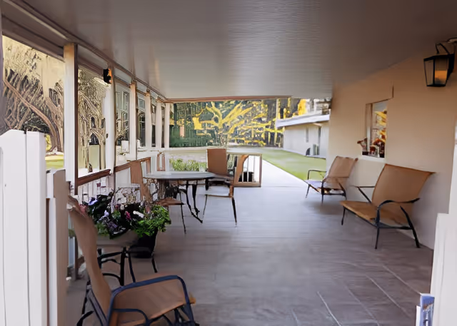Covered outdoor patio area with several chairs and tables arranged along the sides. There are potted plants on the left side near the chairs. The patio overlooks a grassy area and another building is visible in the background.
