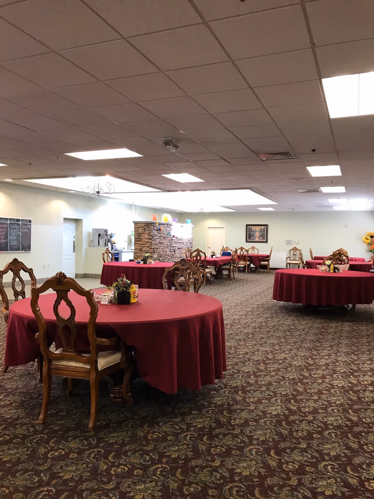 A spacious dining room with several round tables covered in red tablecloths, each surrounded by wooden chairs with cushioned seats. The room has a patterned carpet, a stone partition wall, and bright overhead lighting. There are some decorations on the tables and a chalkboard on the left wall.