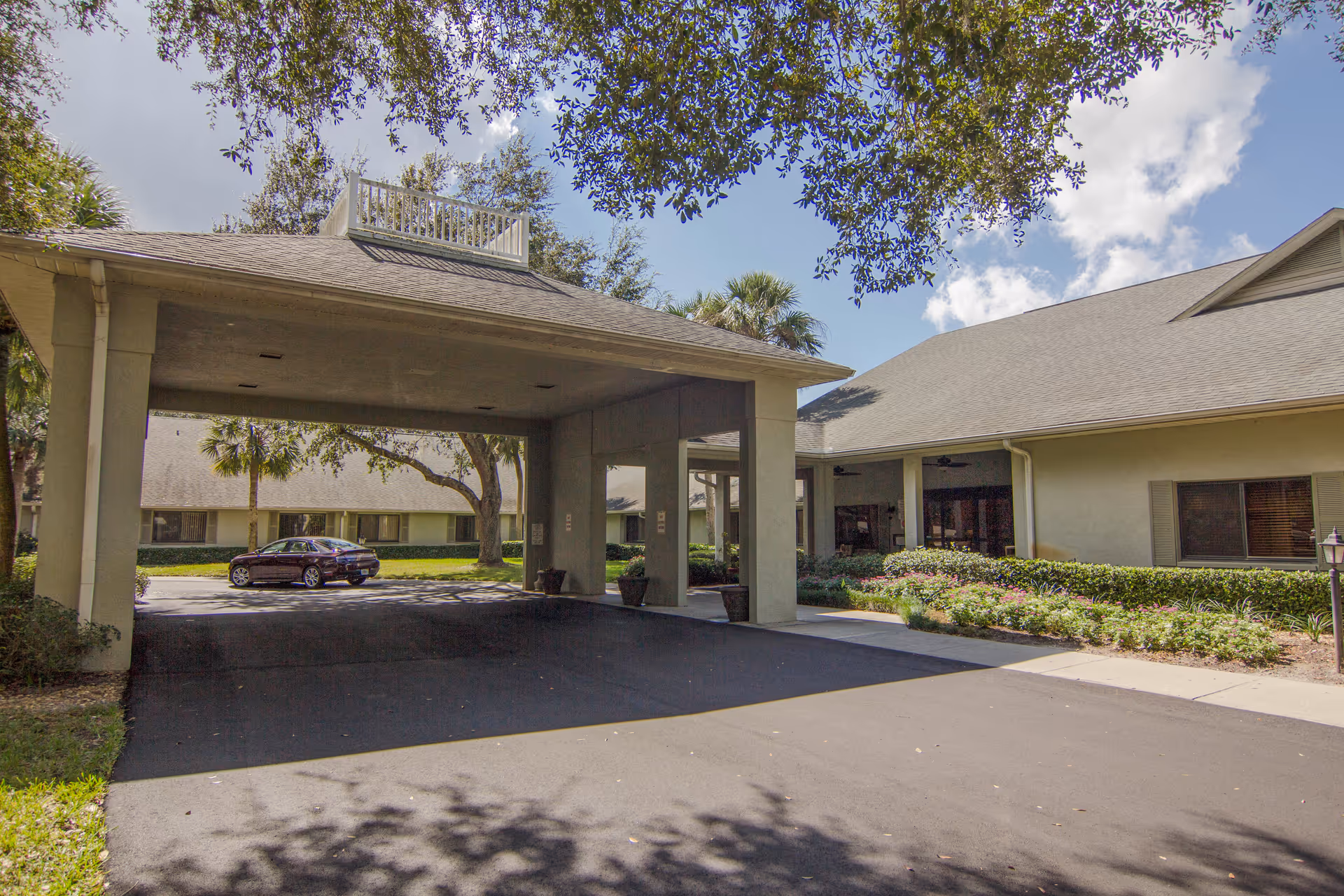 Entrance area of Lake Port Square facility showing a covered driveway with a roof supported by columns, a parked car, surrounding greenery, and parts of the building under a partly cloudy sky.