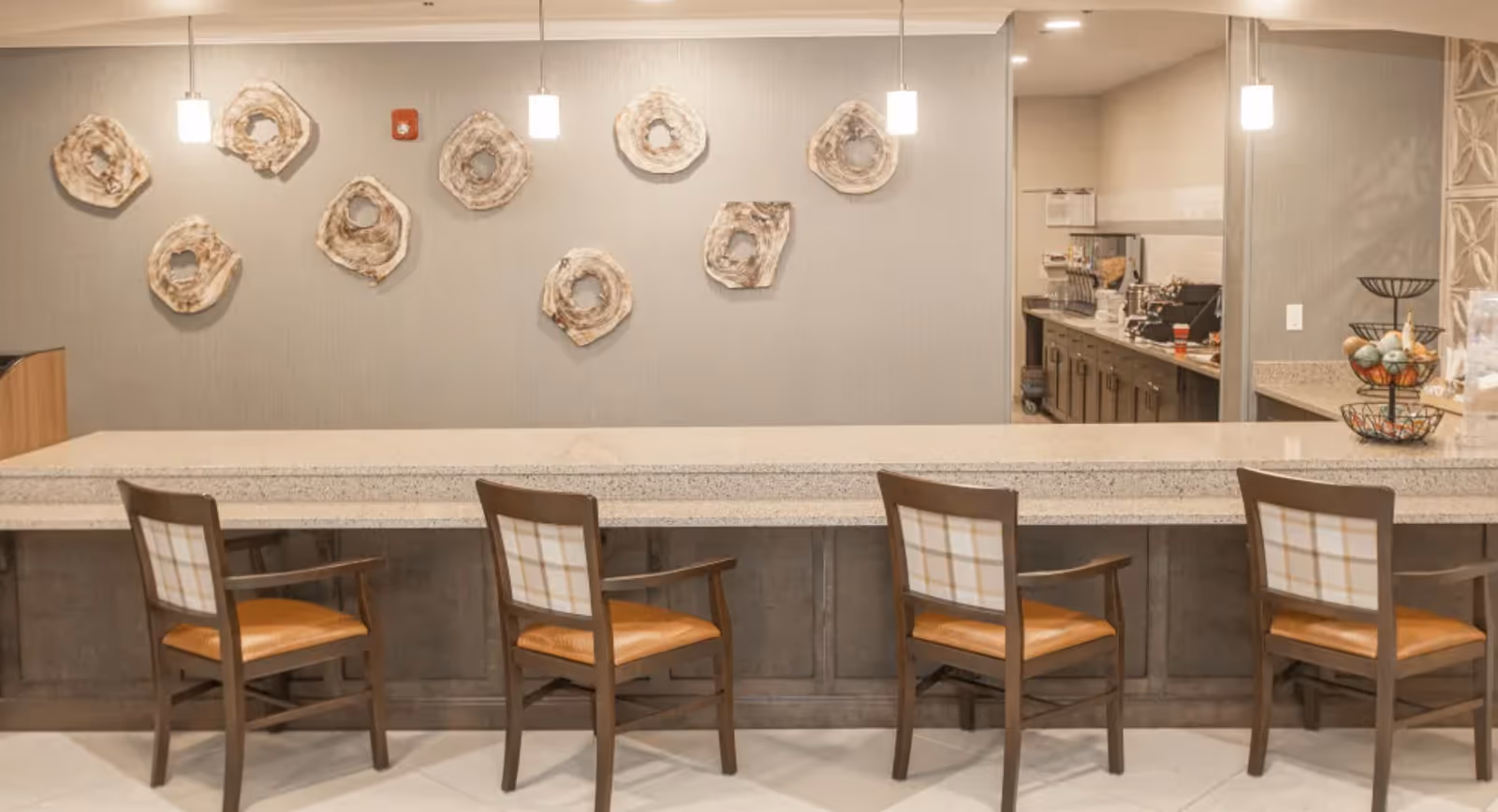 A modern kitchen counter area with four wooden chairs featuring plaid backrests and orange seat cushions. The counter is made of light-colored granite, and the background wall has decorative circular art pieces. Pendant lights hang from the ceiling, and a kitchen area with appliances and cabinets is visible through an open doorway on the right.