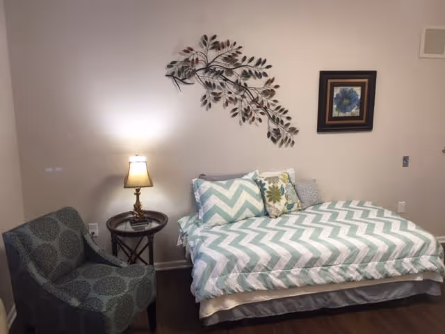 A bedroom with a bed featuring chevron-patterned bedding in light blue and white, multiple decorative pillows, a small round side table with a lamp, a patterned armchair, and wall decorations including a metal leaf sculpture and a framed floral picture.