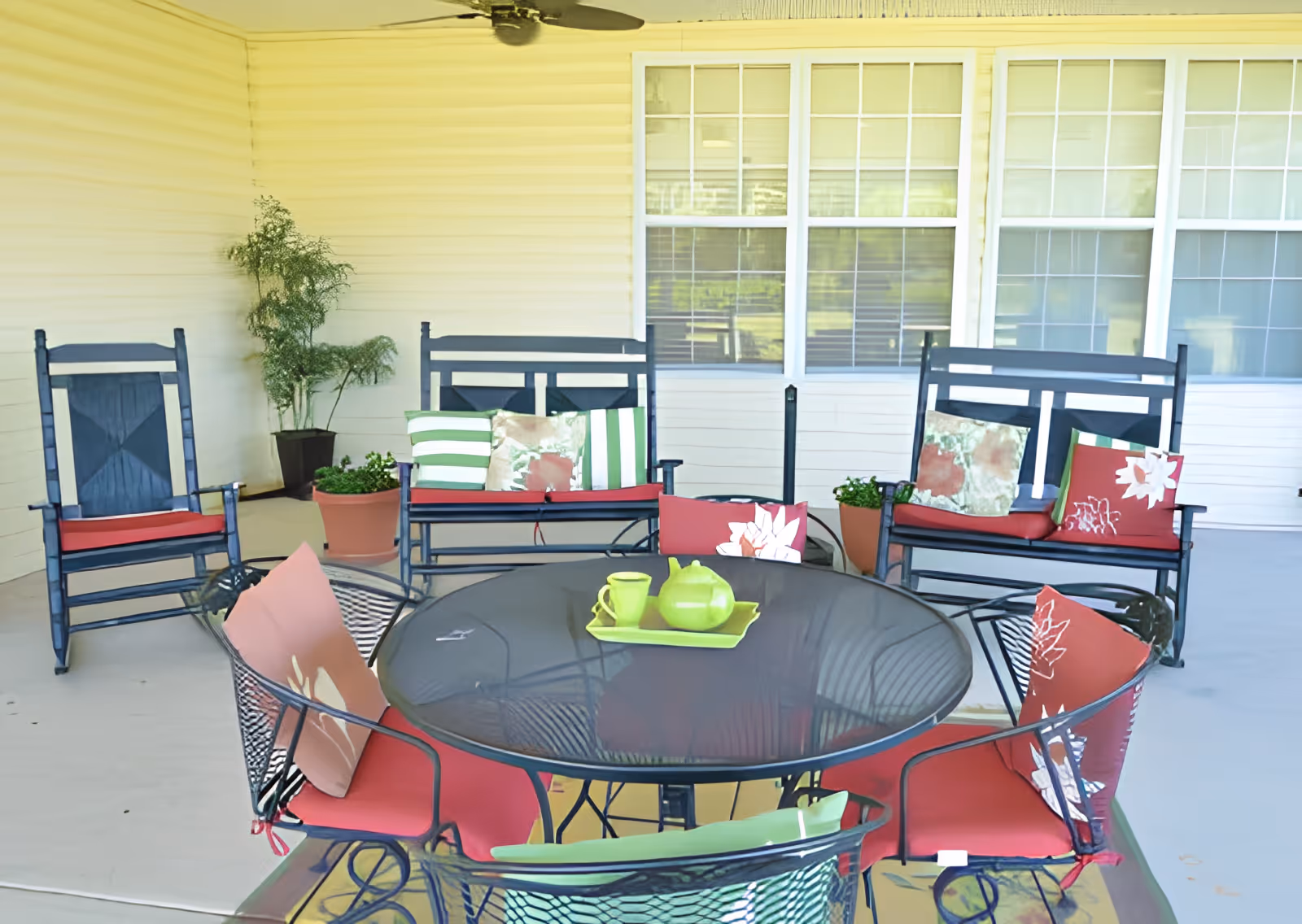 A covered outdoor patio area with black metal chairs and benches featuring red cushions and decorative pillows. A round black metal table with a green teapot and cup set on a tray is in the center. There are potted plants near the walls and large windows in the background.