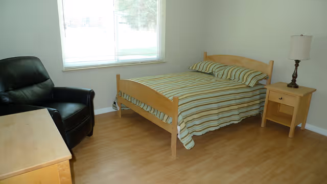 A simple bedroom with a wooden bed frame and striped bedding, a wooden nightstand with a lamp, a black armchair, and a wooden dresser. The room has light-colored walls and a window with blinds letting in natural light.