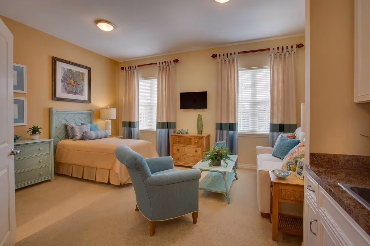 A cozy studio apartment featuring a bed with beige bedding and decorative pillows, a light blue armchair, a white sofa with colorful cushions, a small coffee table with a plant, a wooden dresser with a green vase, two windows with beige and blue curtains, and a wall-mounted TV. The room has warm beige walls and carpeted flooring.