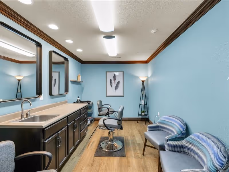 Interior of a salon room with light blue walls and wooden floor. The room features two salon chairs in front of a long counter with a sink and two large mirrors. There are two patterned chairs along the right wall and a tall floor lamp in the corner. A framed picture of feathers hangs on the back wall.
