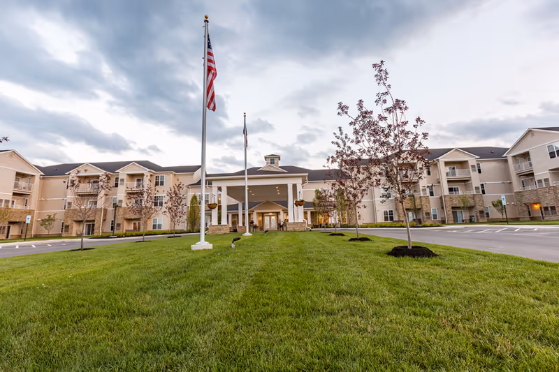 Exterior view of StoryPoint Troy senior living facility showing a large, three-story building with balconies, a covered entrance with columns, two flagpoles with American and state flags, and a well-maintained lawn with young trees under a cloudy sky.