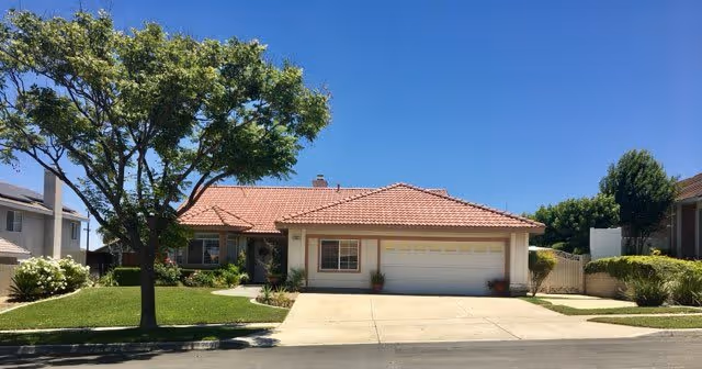 Single-story house with a red tile roof, a two-car garage, and a well-maintained front yard with a large tree and green grass under a clear blue sky.