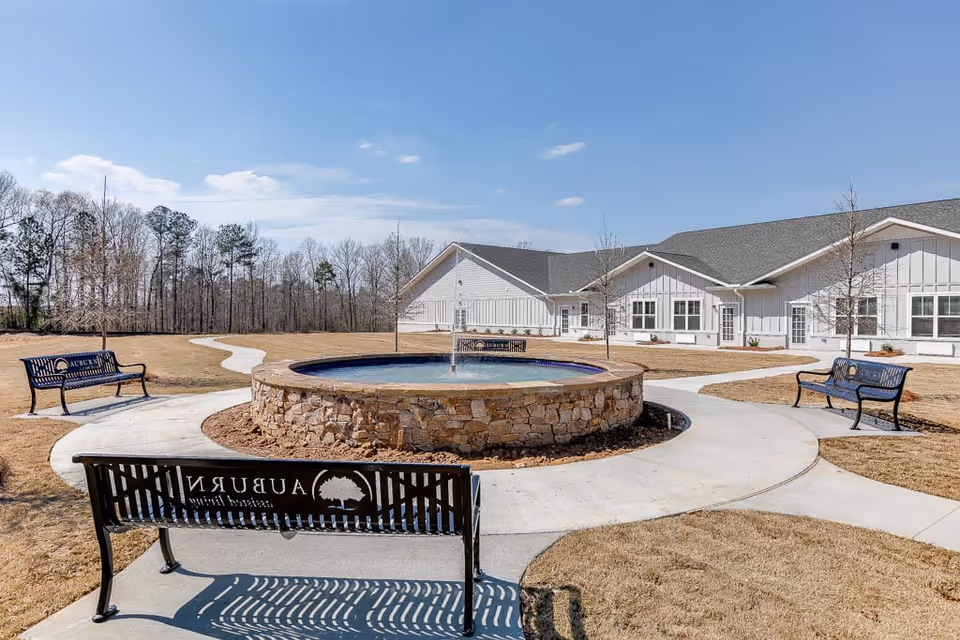 Outdoor courtyard area at Auburn Assisted Living featuring a circular stone fountain in the center surrounded by concrete pathways and three black metal benches with Auburn Assisted Living logos. The building is visible in the background under a clear blue sky with some trees beyond the courtyard.
