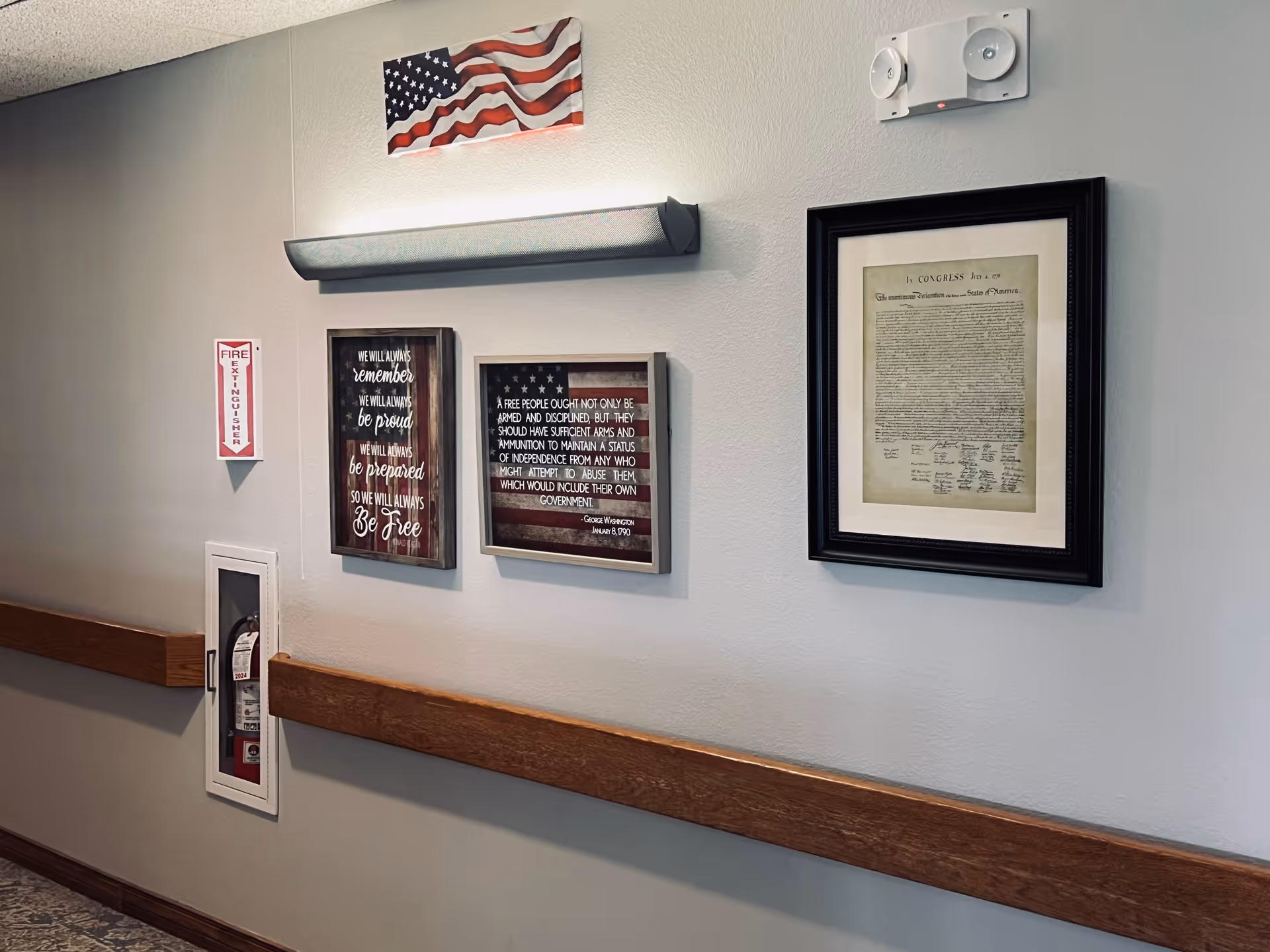Hallway wall with framed patriotic artwork and a framed document above a fire extinguisher cabinet and wooden handrail.