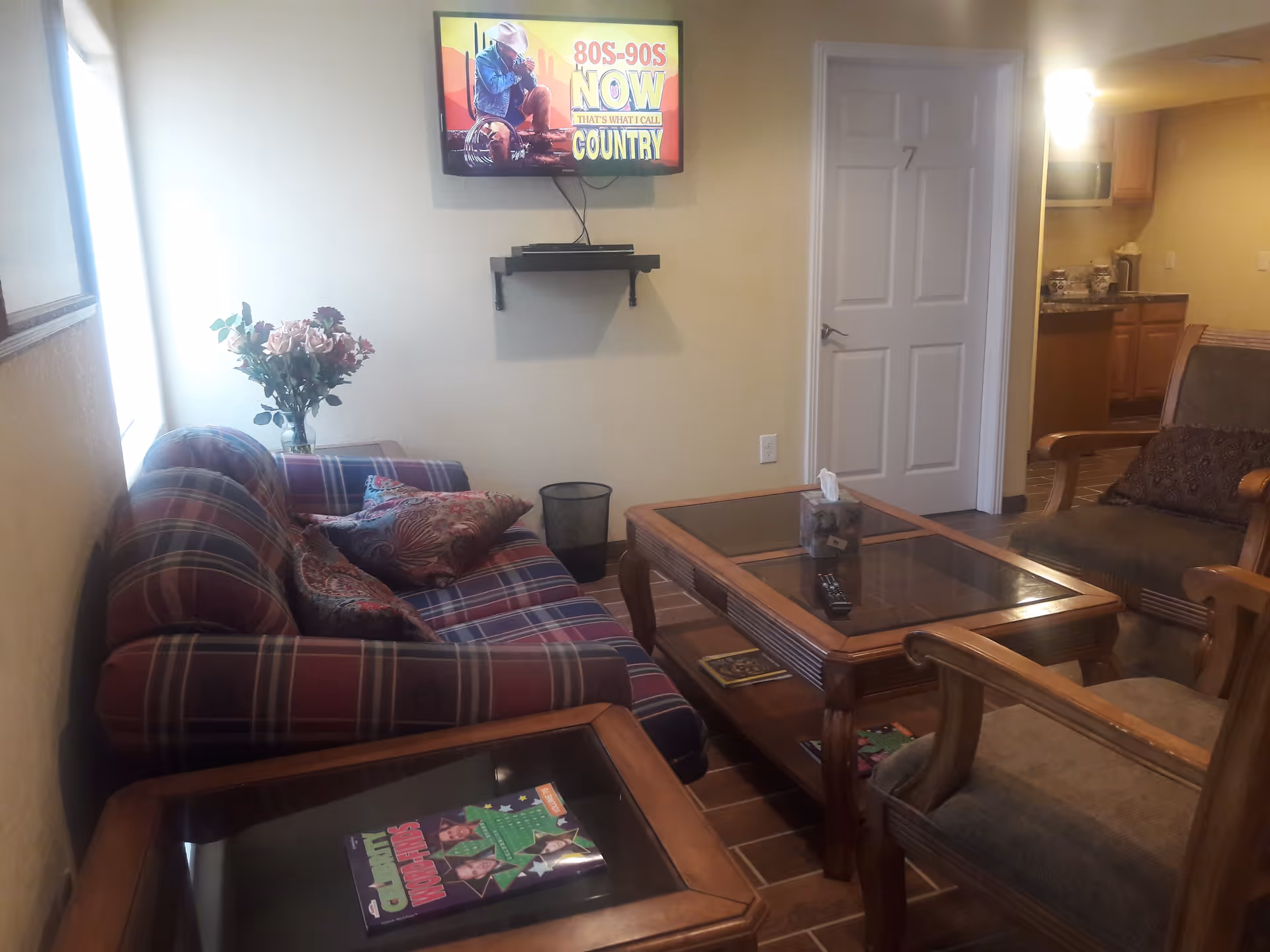 A cozy living room area with a plaid sofa, two cushioned armchairs, a wooden coffee table with glass top, and a side table with magazines. A flat-screen TV is mounted on the wall showing a country music channel. There is a vase with flowers on a small table near a window, and a door labeled with the number 7 leads to another room. The background shows a kitchen area with wooden cabinets and a microwave.
