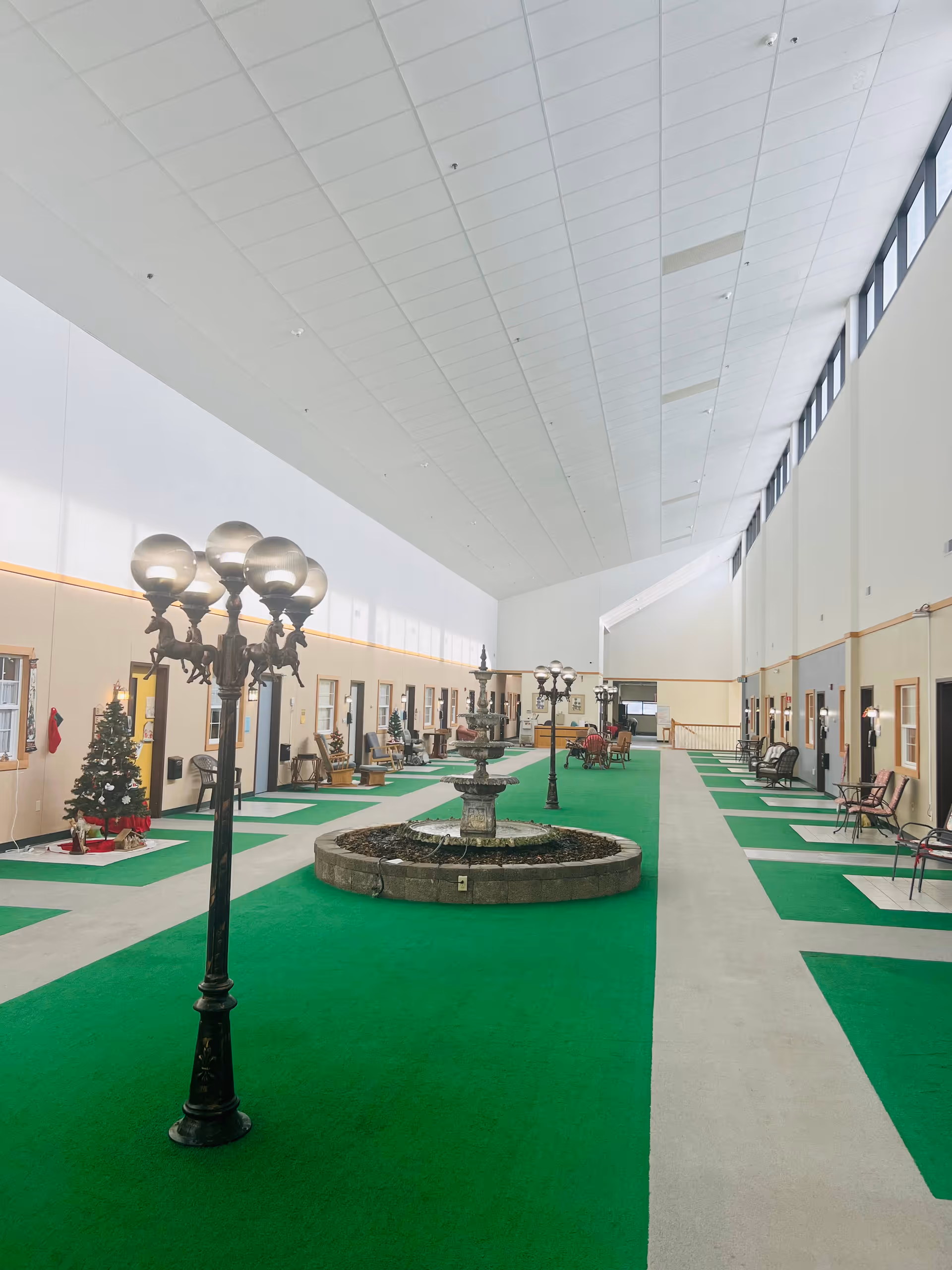 Indoor courtyard area with green carpet flooring and a central stone fountain. The space is lined with doors and windows on both sides, each with chairs and small tables outside. Decorative streetlamp-style lights and a Christmas tree are visible. The ceiling is high with windows near the top allowing natural light.