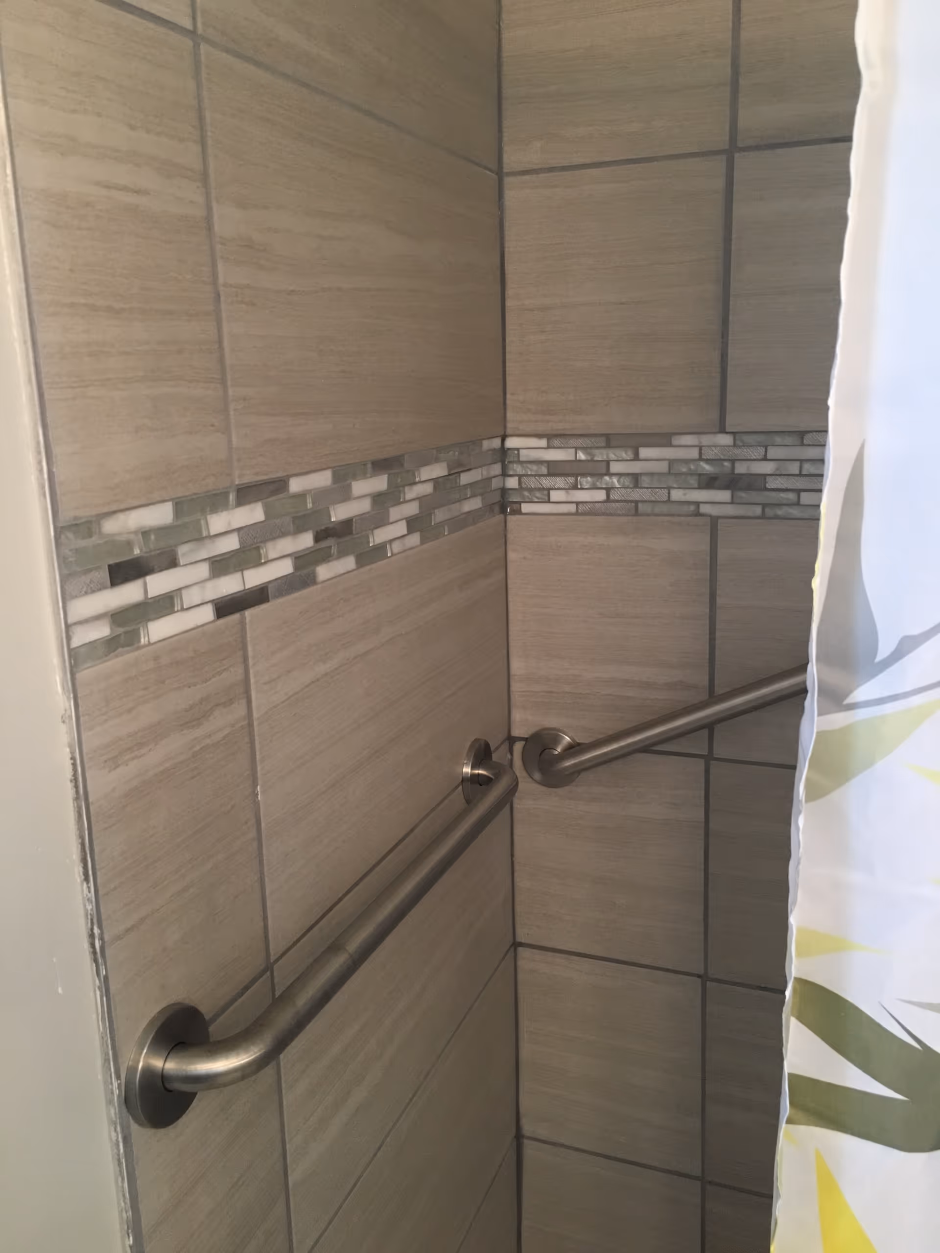 Corner of a shower area with beige and gray tiled walls, featuring a horizontal decorative strip of small rectangular tiles in shades of white, gray, and green. There are two stainless steel grab bars mounted on the walls for support, and a partially visible shower curtain with a green and yellow leaf pattern on the right side.