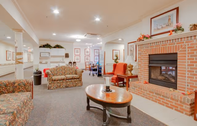 A cozy senior living facility common area with floral patterned sofas, a wooden coffee table, a brick fireplace decorated with framed pictures and flowers, and a small kitchenette in the background. The room is well-lit with ceiling lights and has carpeted flooring.