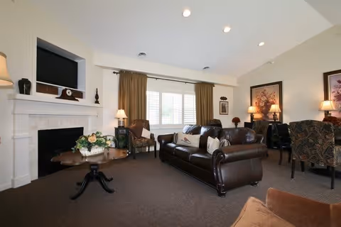 A cozy living room with a dark brown leather sofa, two patterned armchairs, a wooden coffee table with a floral arrangement, a fireplace with a TV mounted above it, and a window with brown curtains letting in natural light. The room also features several lamps and framed artwork on the walls.