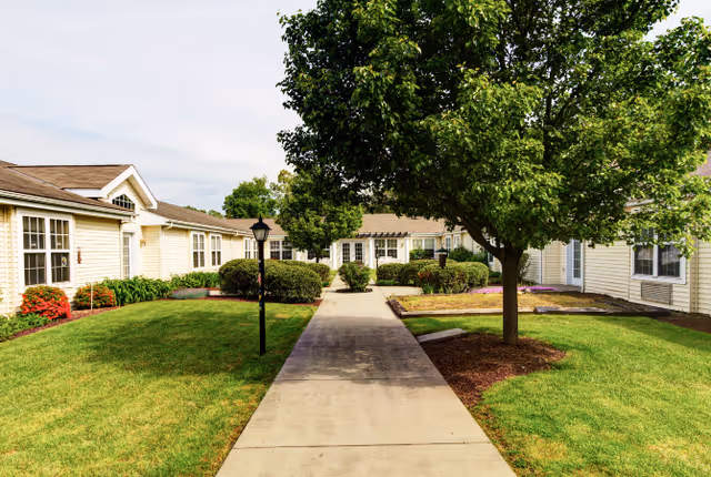 A paved walkway leading through a well-maintained garden area with green grass, bushes, and a large tree, surrounded by single-story beige buildings with white-framed windows under a partly cloudy sky.
