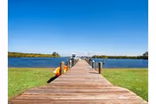 A long wooden dock extending over a body of water under a clear blue sky, with green grass on either side of the dock and a small orange kayak resting on the left side near the water.