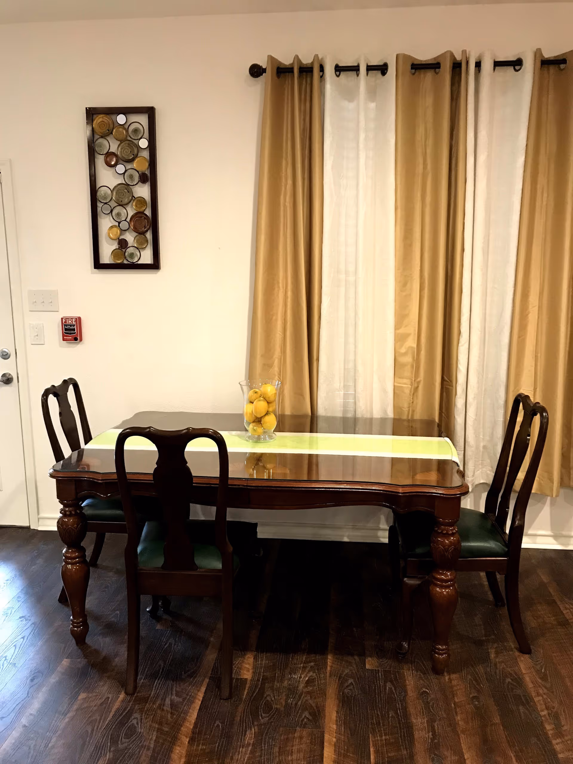 A dining area with a wooden table and four matching chairs with dark green cushions. The table has a glass top with a light green runner and a glass vase filled with lemons. Behind the table are beige and white curtains hanging on a black curtain rod. On the left wall, there is a decorative wall art piece with circular designs and a red fire alarm.