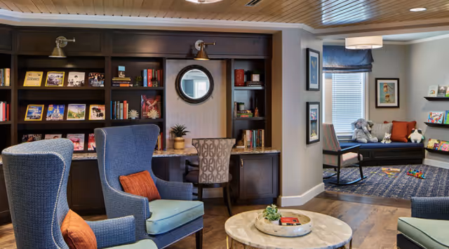 A cozy interior common area with dark wood bookshelves filled with books and magazines, two blue upholstered armchairs with orange pillows, a round marble coffee table with a decorative tray, a desk with a patterned chair and a round mirror above it, and a small play area with a rocking chair, stuffed animals, and children's books near a window with blue curtains.