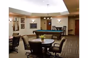 Interior view of a common area in a senior living facility named Prairie View, featuring a pool table in the center, round tables with chairs around them, carpeted floor, wall-mounted lights, and framed pictures on the walls.
