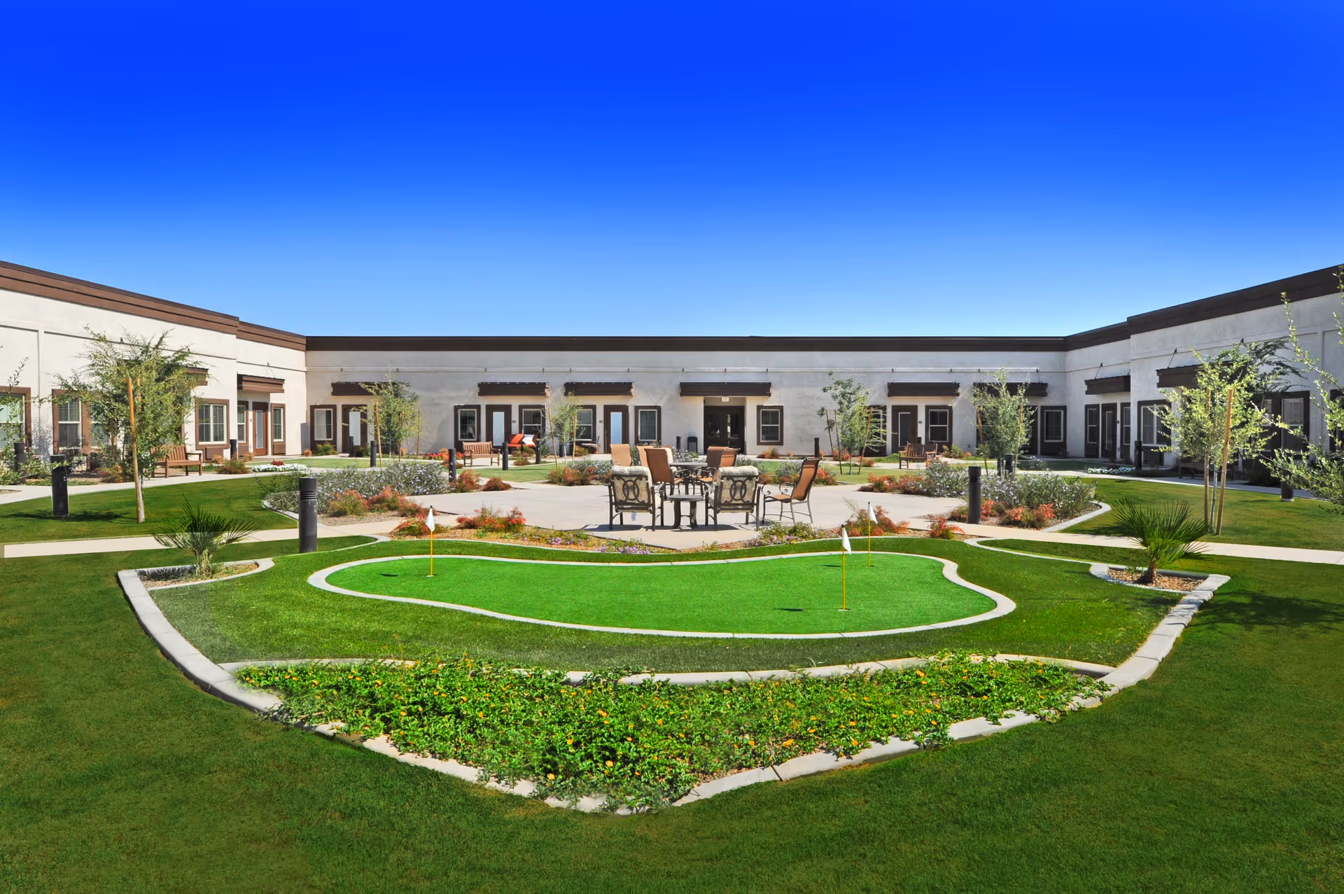 Outdoor courtyard area of a senior living facility with a small putting green, patio table and chairs, landscaped garden beds, and surrounding single-story building with multiple doors and windows under a clear blue sky.