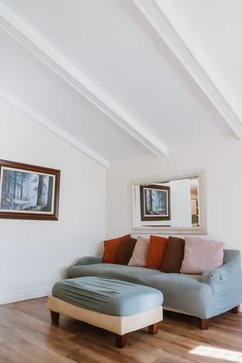 Small living room with a light blue sofa topped with colorful throw pillows, a matching ottoman, a wall mirror and framed artwork beneath a white beamed ceiling.