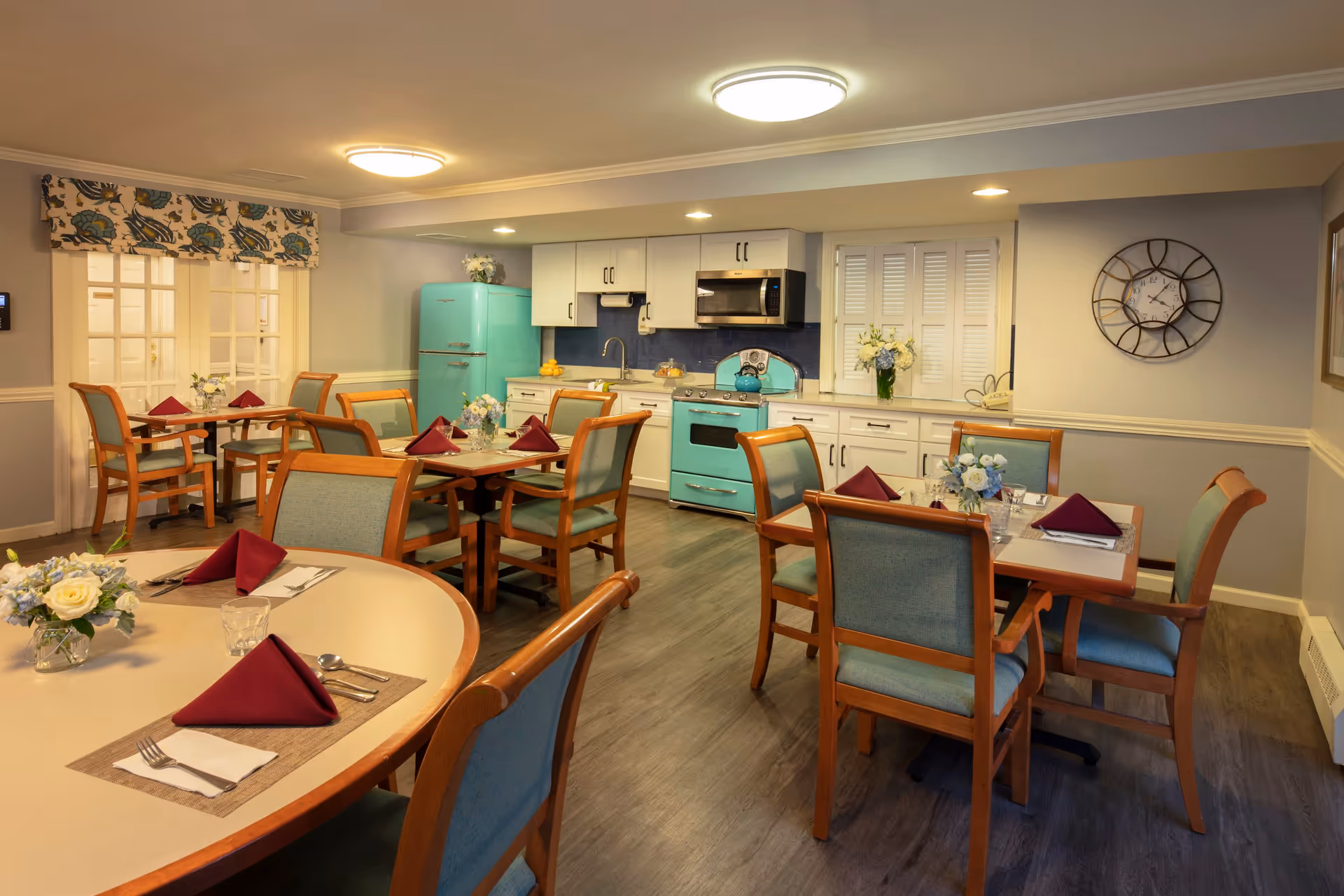A cozy dining room with several wooden tables and chairs set with burgundy napkins, silverware, and floral centerpieces. In the background, there is a kitchen area with white cabinets, a turquoise retro-style refrigerator and stove, a microwave, and a sink. The room has soft lighting from ceiling fixtures and a wall clock is visible on the right wall.