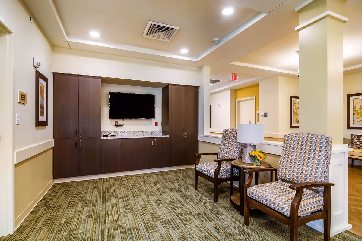 A well-lit interior common area with two patterned armchairs and a small round table with a lamp and a small flower arrangement. Behind the chairs is a half wall with a column. On the opposite wall, there is a mounted flat-screen TV above a long cabinet with dark wood doors and a granite countertop. The room has light-colored walls, framed artwork, and a green patterned carpet.