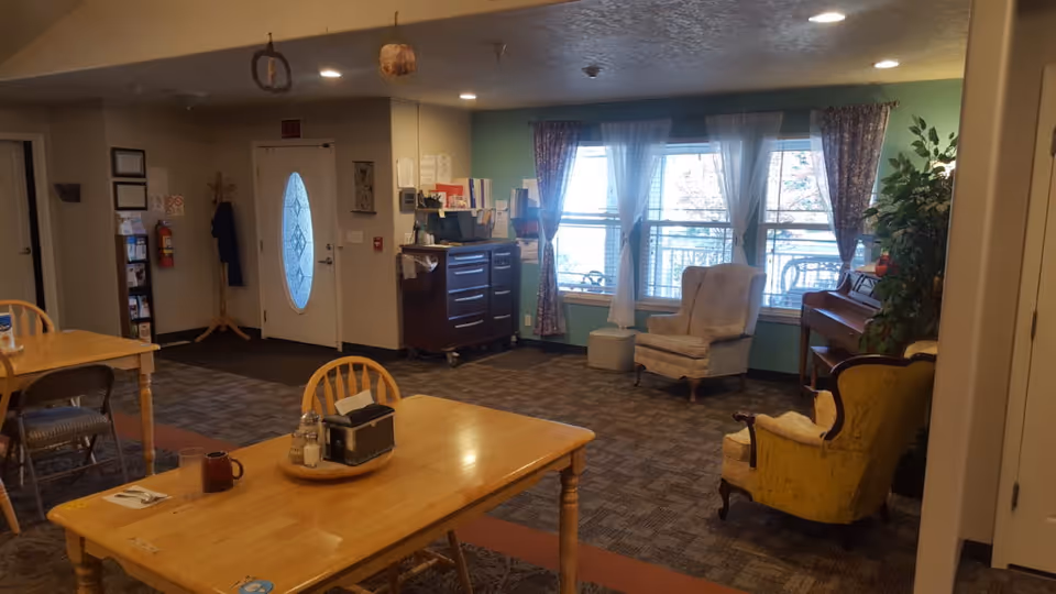 A senior living common room with wooden dining tables and chairs in the foreground, upholstered armchairs and a piano by the windows, and an entrance door.