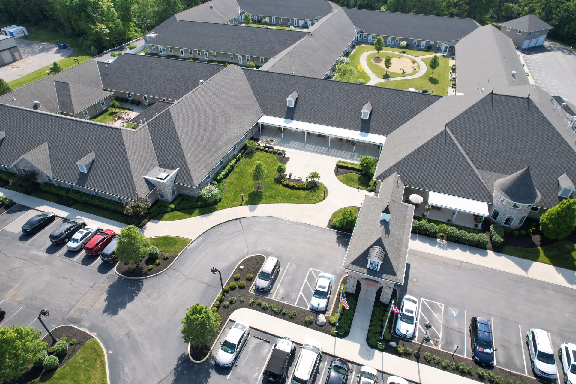 Aerial view of Hudson Grande Senior Living facility showing multiple connected buildings with dark roofs surrounding landscaped courtyards with green lawns, trees, and walking paths. There is a parking lot with several cars and a small entrance structure with flags near the front.