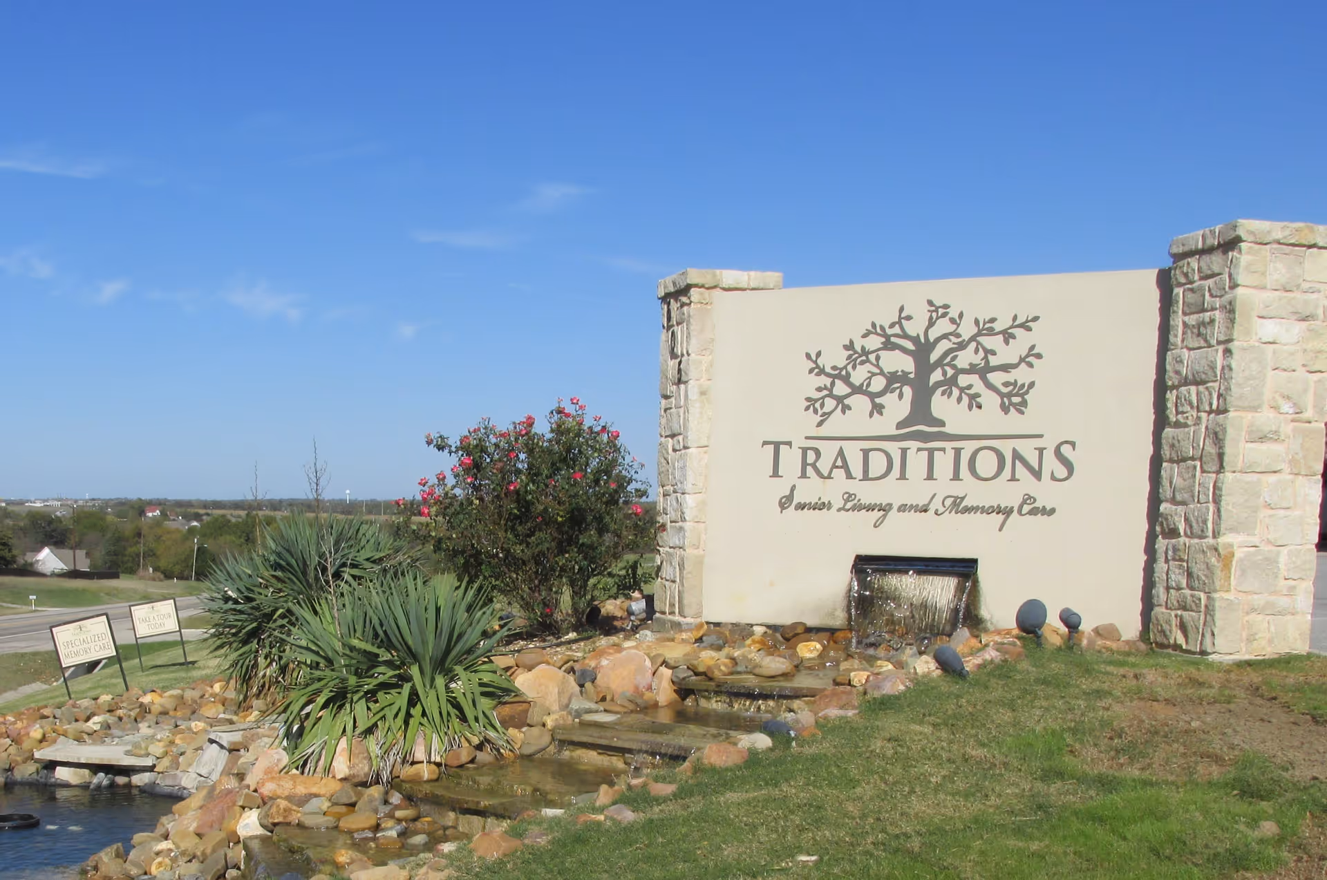 Outdoor view of a stone and stucco sign for Traditions Senior Living and Memory Care, featuring a tree logo and a small water feature with rocks and plants around it under a clear blue sky.