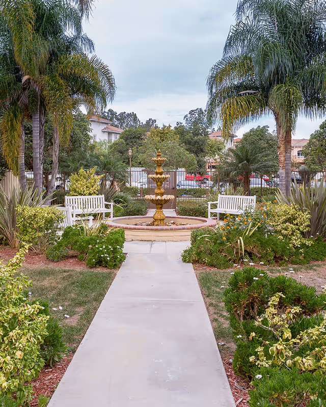 A garden pathway leading to a decorative multi-tiered fountain surrounded by greenery, palm trees, and two white benches on either side. Residential buildings and a parking lot are visible in the background under a cloudy sky.