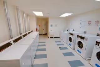 A bright laundry room with multiple white washing machines and dryers lined up along the walls. The floor has a checkered pattern with light and dark tiles, and there are fluorescent lights on the ceiling. The room appears clean and well-lit.
