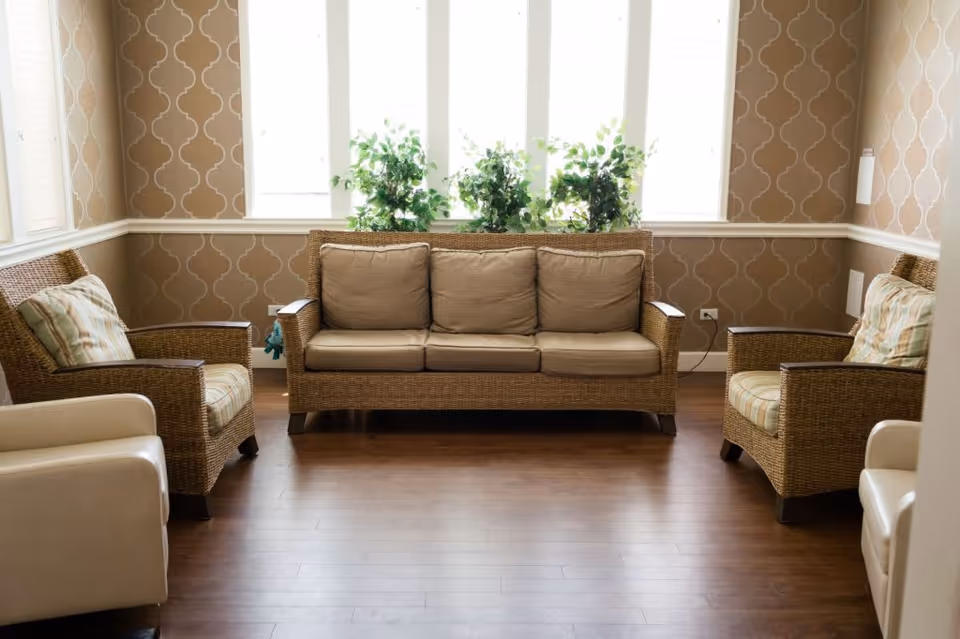 A cozy sitting area with a wicker sofa and two wicker armchairs with cushions, arranged around a wooden floor. The walls have a patterned wallpaper, and there are large windows behind the sofa letting in natural light, with green plants placed on the windowsill.