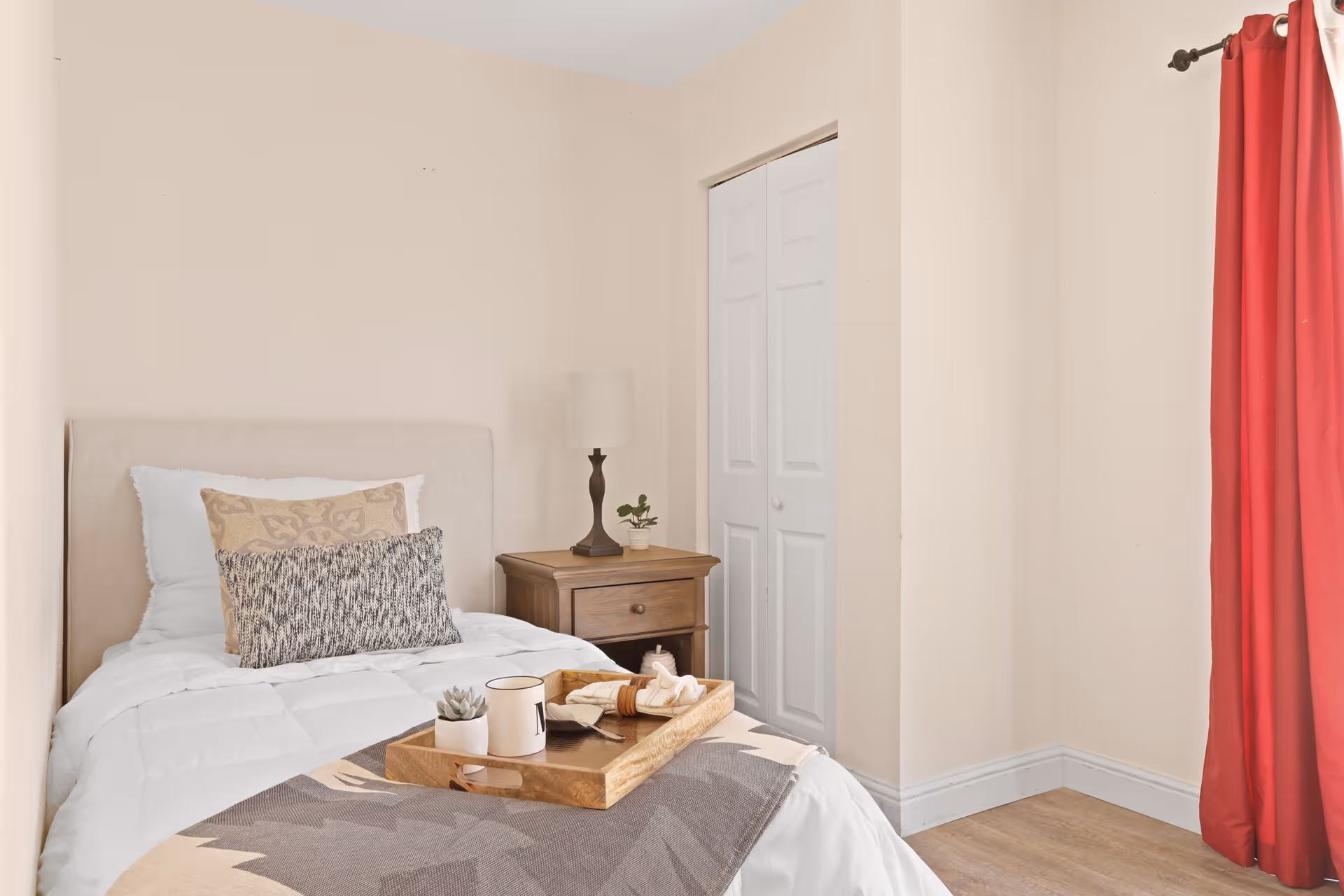 A cozy bedroom with a single bed featuring white bedding and multiple pillows. A wooden nightstand with a lamp and a small plant is next to the bed. There is a closed white closet door on the wall and a window with red curtains on the right side.