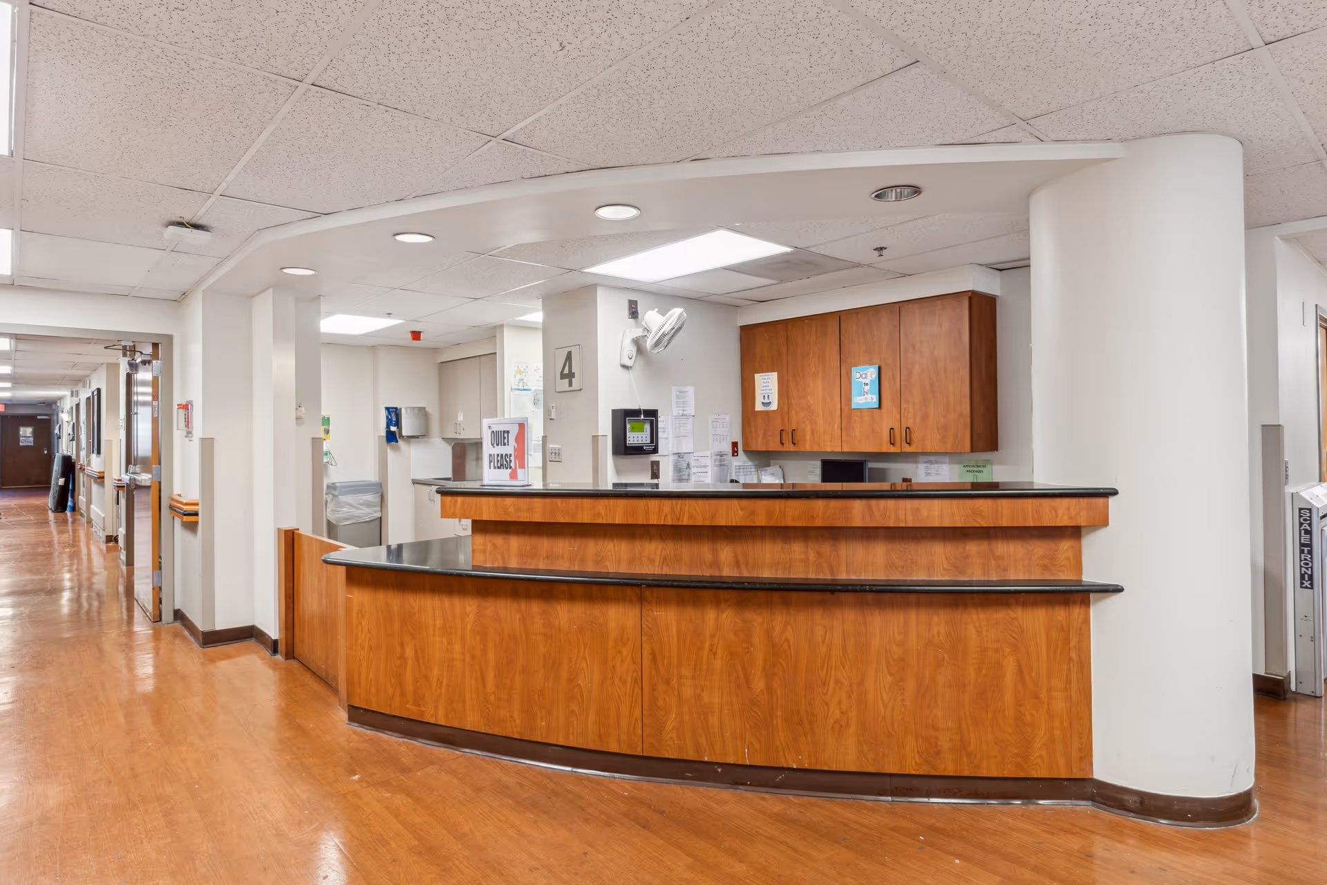 Nursing station/reception area inside a senior care facility hallway with a curved wooden desk and overhead cabinets.