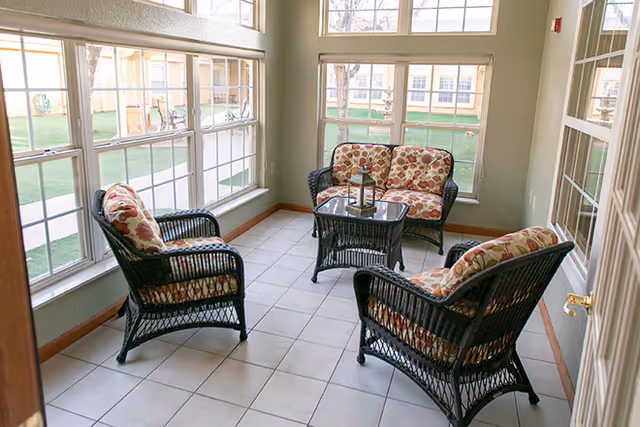 A sunlit indoor seating area with large windows on two sides, featuring four black wicker chairs with floral cushions arranged around a small glass-top wicker table. The floor is tiled, and the room has light-colored walls with wooden trim.