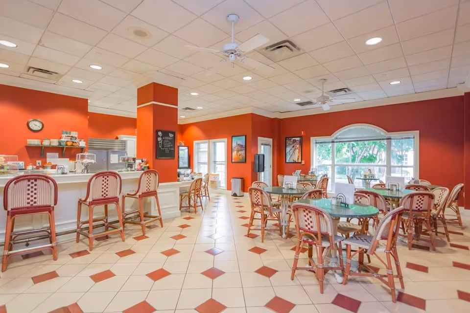A bright dining area with red walls and white tiled floor featuring red diamond patterns. There are several tables with chairs arranged around them, and a counter with bar stools on the left side. Large windows allow natural light to enter the room, and ceiling fans hang from the white ceiling.