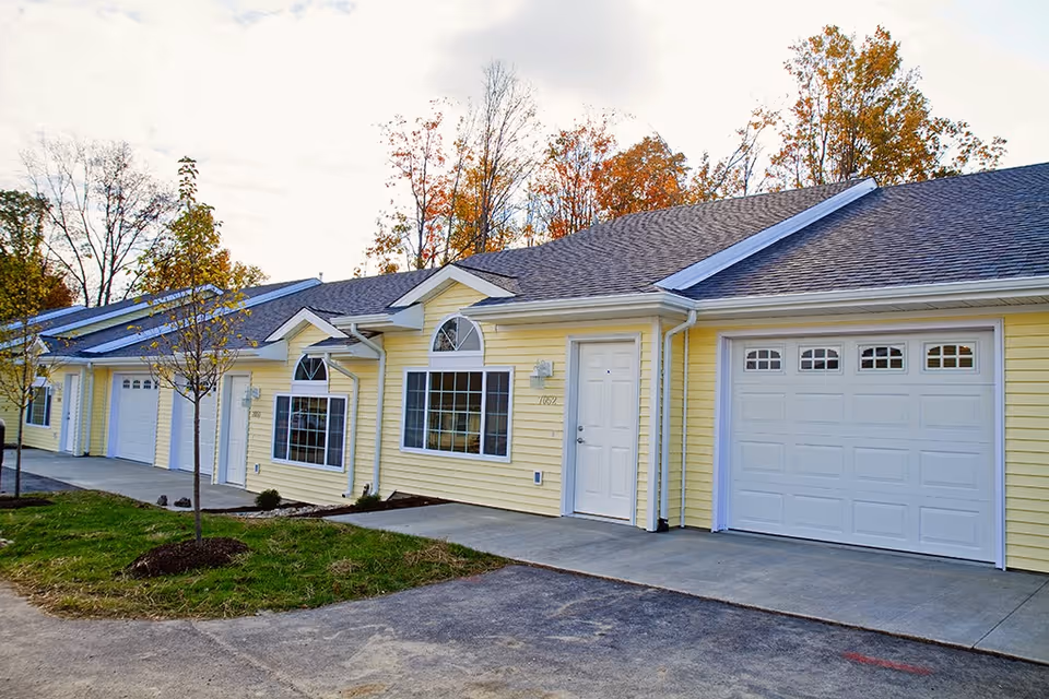 Exterior view of a single-story yellow retirement community building with multiple units, each featuring a white garage door, a white entrance door, and windows. Small trees and landscaping are visible in front of the building, with autumn-colored trees in the background.