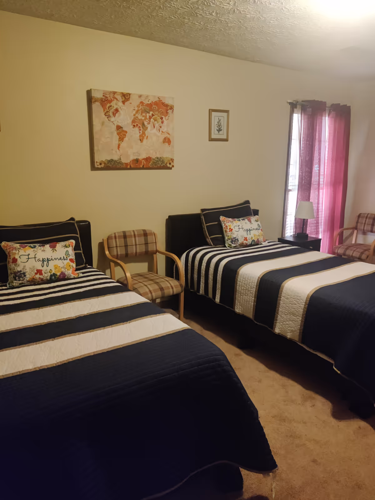 A bedroom with two twin beds covered in navy blue and white striped bedding. Each bed has a decorative pillow with the word 'Happiness' and floral designs. Between the beds is a plaid armchair, and another matching armchair is near a window with purple curtains. The walls are beige with two framed pictures, one of a world map and one of a plant.