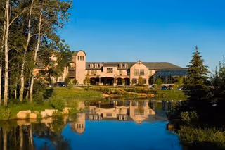 A large, multi-story building with stone and brick exterior situated behind a pond reflecting the building and surrounding trees under a clear blue sky.