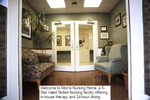 Entrance area of Morris Nursing Home featuring a floral patterned sofa on the left and a light blue armchair on the right, with potted plants and framed pictures on the walls. Double glass doors lead to a hallway inside the facility.