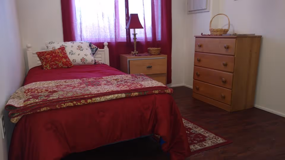 A cozy bedroom with a single bed covered in a red bedspread and floral pillows. Next to the bed is a wooden nightstand with a red lampshade and a small basket. Across from the bed is a wooden dresser with four drawers and a basket on top. The room has dark wood flooring and a window with red curtains letting in soft light.