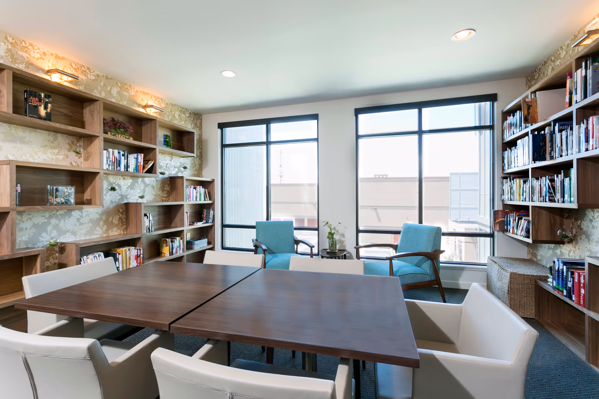 A bright reading or meeting room with large windows letting in natural light. The room features wooden bookshelves filled with books along two walls, a dark wooden table with white cushioned chairs around it, and two blue armchairs near the windows with a small round table between them holding a small plant.