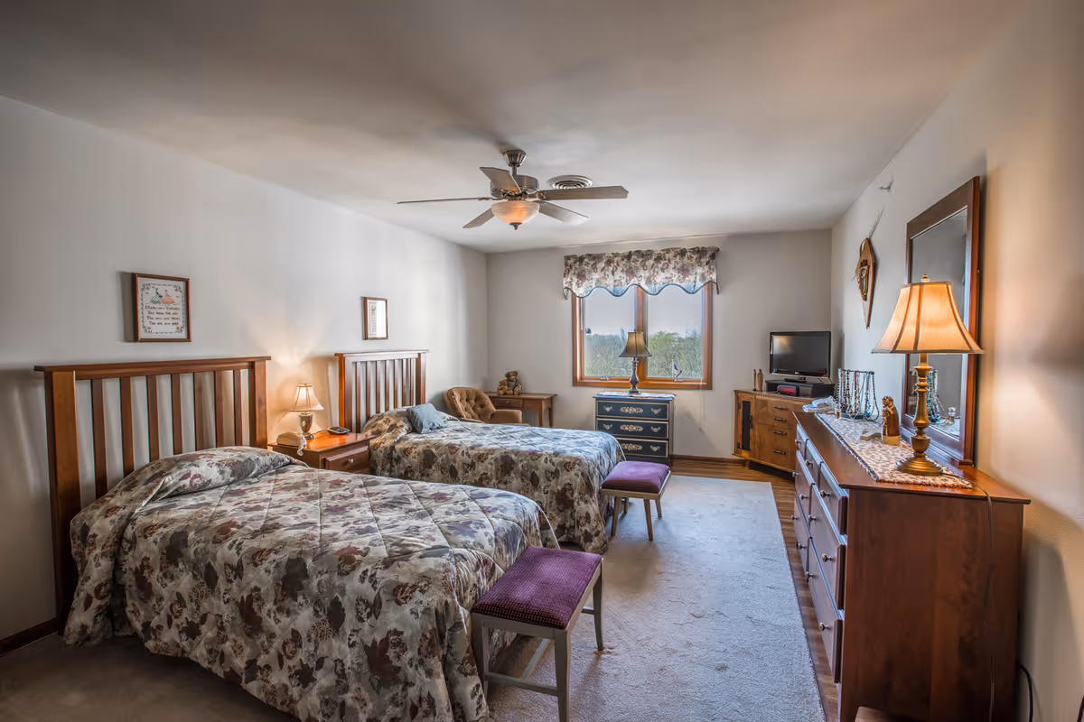 A bedroom with two twin beds featuring floral bedspreads, each with a wooden headboard. Between the beds is a nightstand with a lamp and a phone. At the foot of each bed is a small bench with a purple cushion. The room has a ceiling fan with a light, a window with floral valance, a dresser with a lamp and mirror, a small TV on a wooden cabinet, and a cozy armchair in the corner.