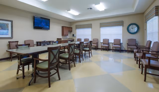 A spacious room with multiple chairs arranged along the walls and around two rectangular tables in the center. The room has large windows with blinds, a wall-mounted TV, a clock, and a small kitchenette area with cabinets and a countertop. The floor has a checkered pattern in beige and light gray tones.